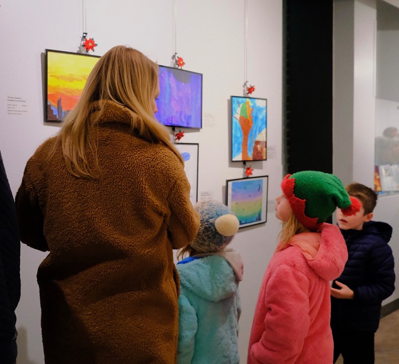 A family of one parent and three children standing in front of holiday-themed artwork on a gallery wall in conversation
