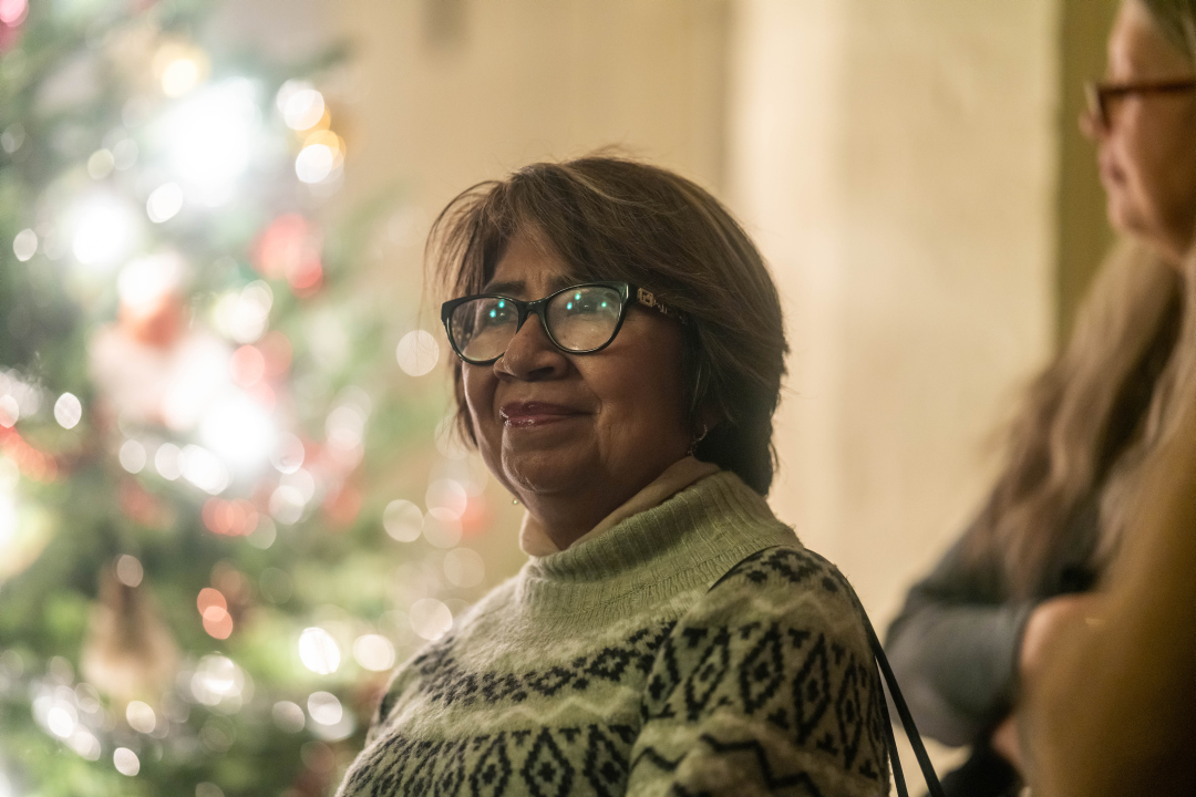 Twinkling christmas lights are reflected in the eyeglasses of a woman dressed in a cozy sweater.