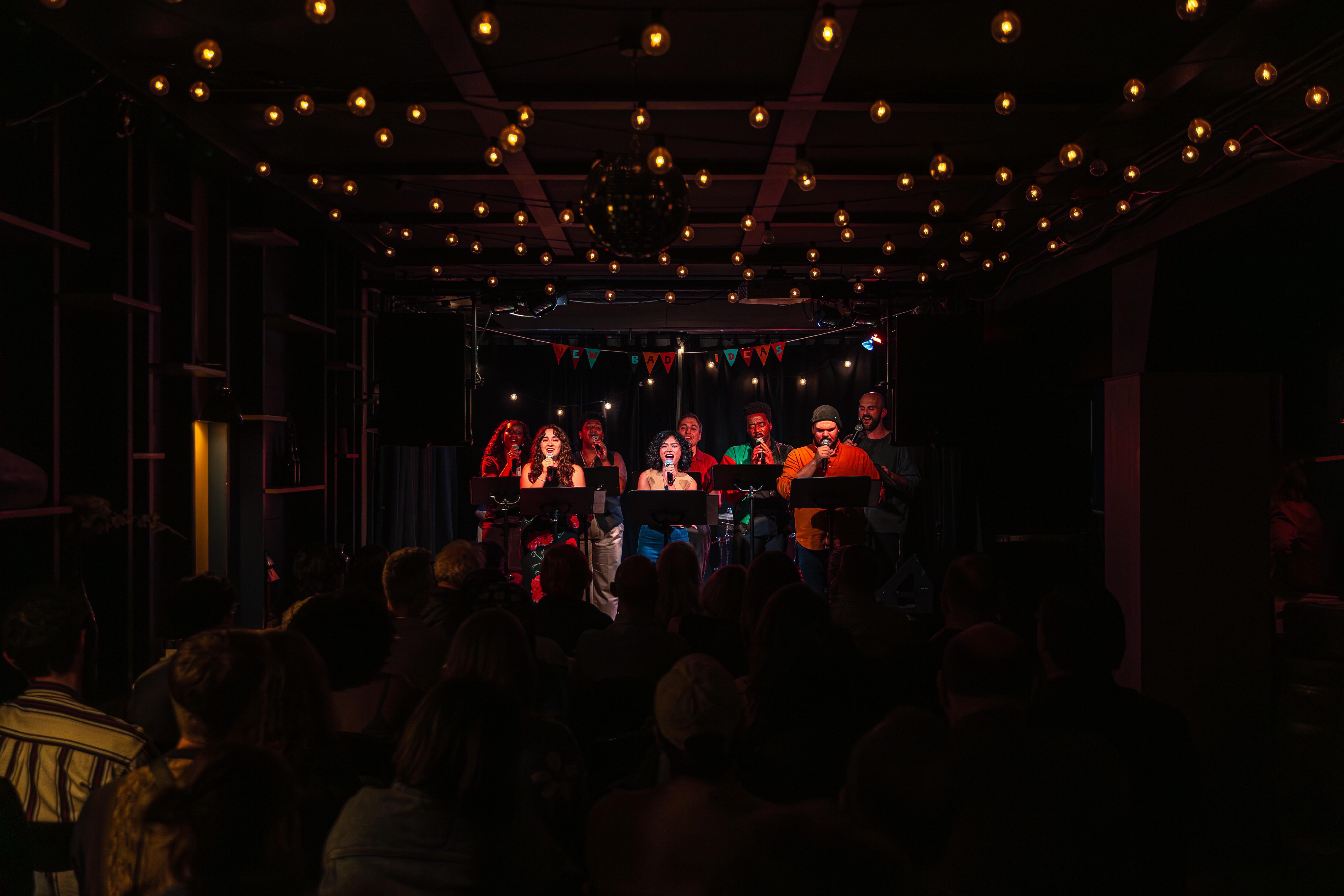 Eight performers stand on a well-lit stage with microphones in hand.