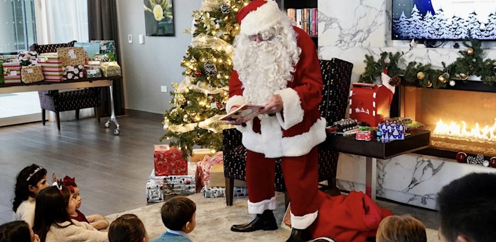 Santa Claus reads to a group of children gathered around a decorated Christmas tree, surrounded by wrapped presents and