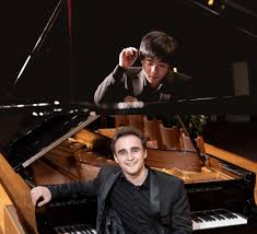 Illia Ovcharenko, leans in front of a grand piano. Kevin Chen sits down in front of the keys.