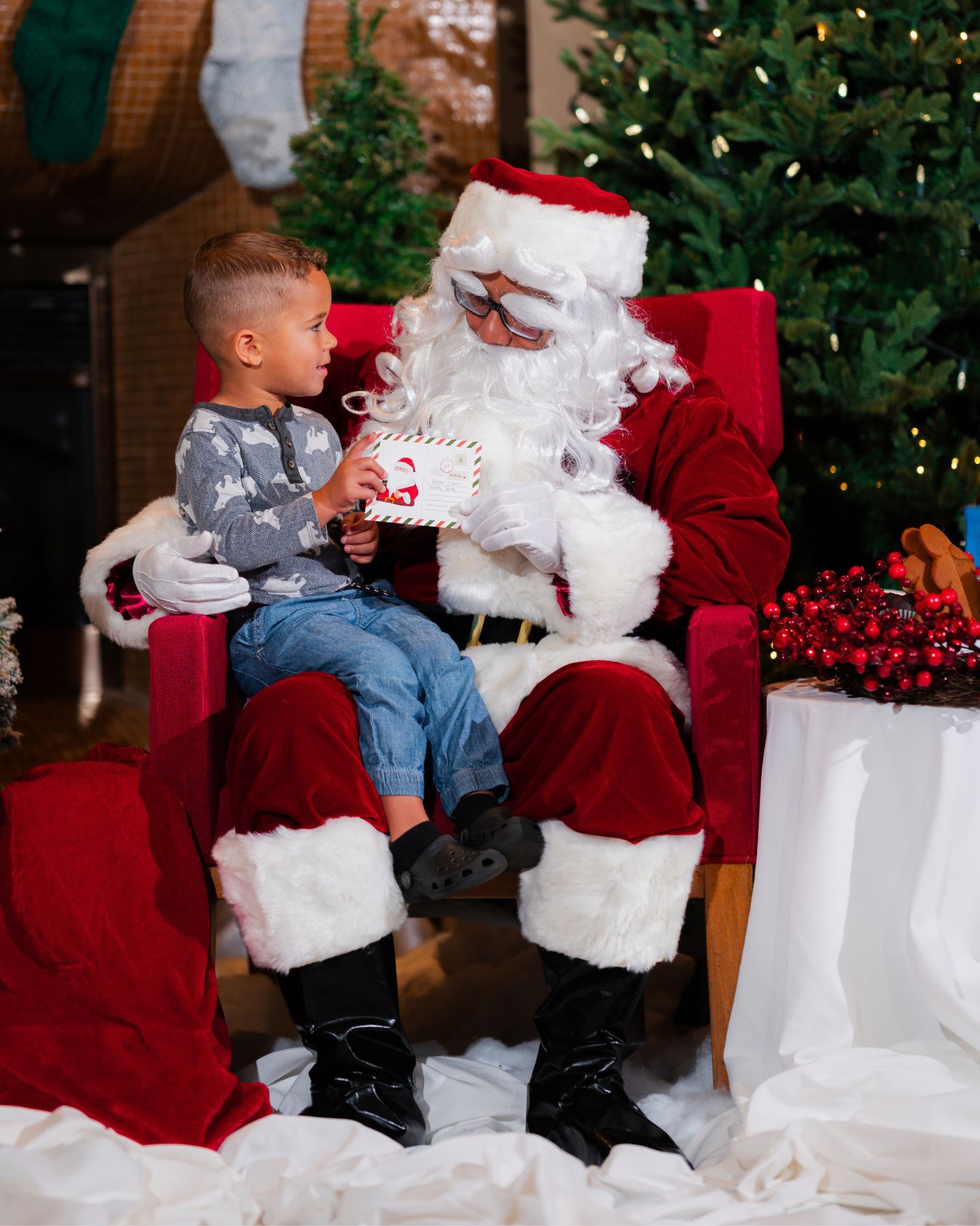 Man dressed as Santa Claus sits on a red chair with a young child on his lap, holding a letter.