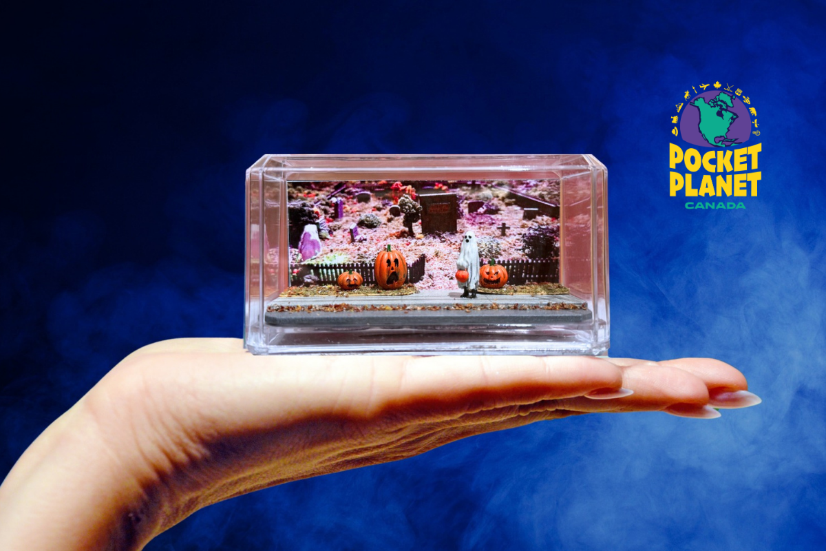 Miniature Halloween graveyard scene with pumpkins, a ghost and tombstones inside a clear display case, resting on hand
