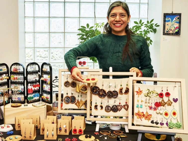 Woman  wearing a green sweater stands behind her handmade jewelry stand.