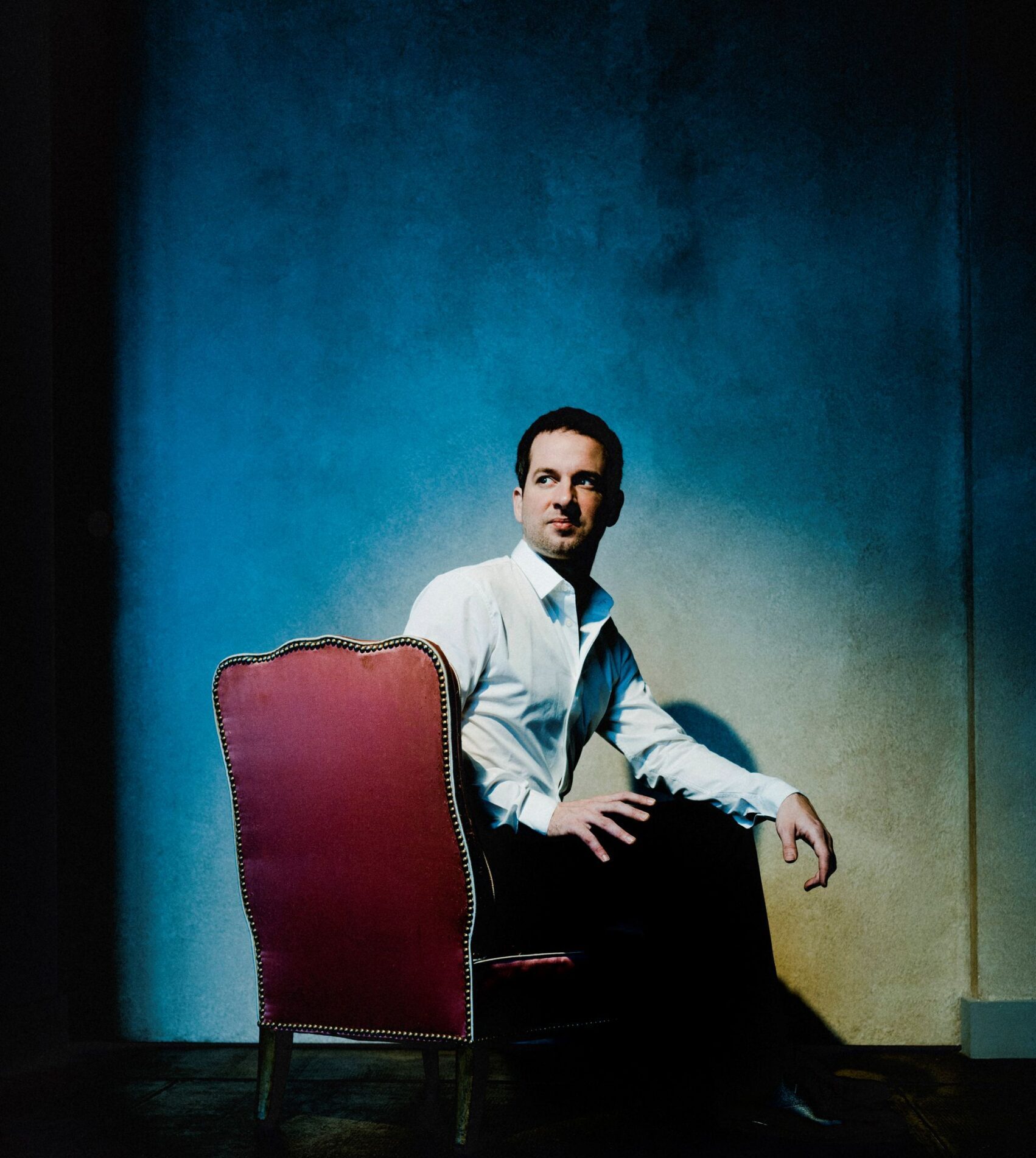 Bertrand Chamayou sits on a red chair with a spotlight shinning at him over a blue background