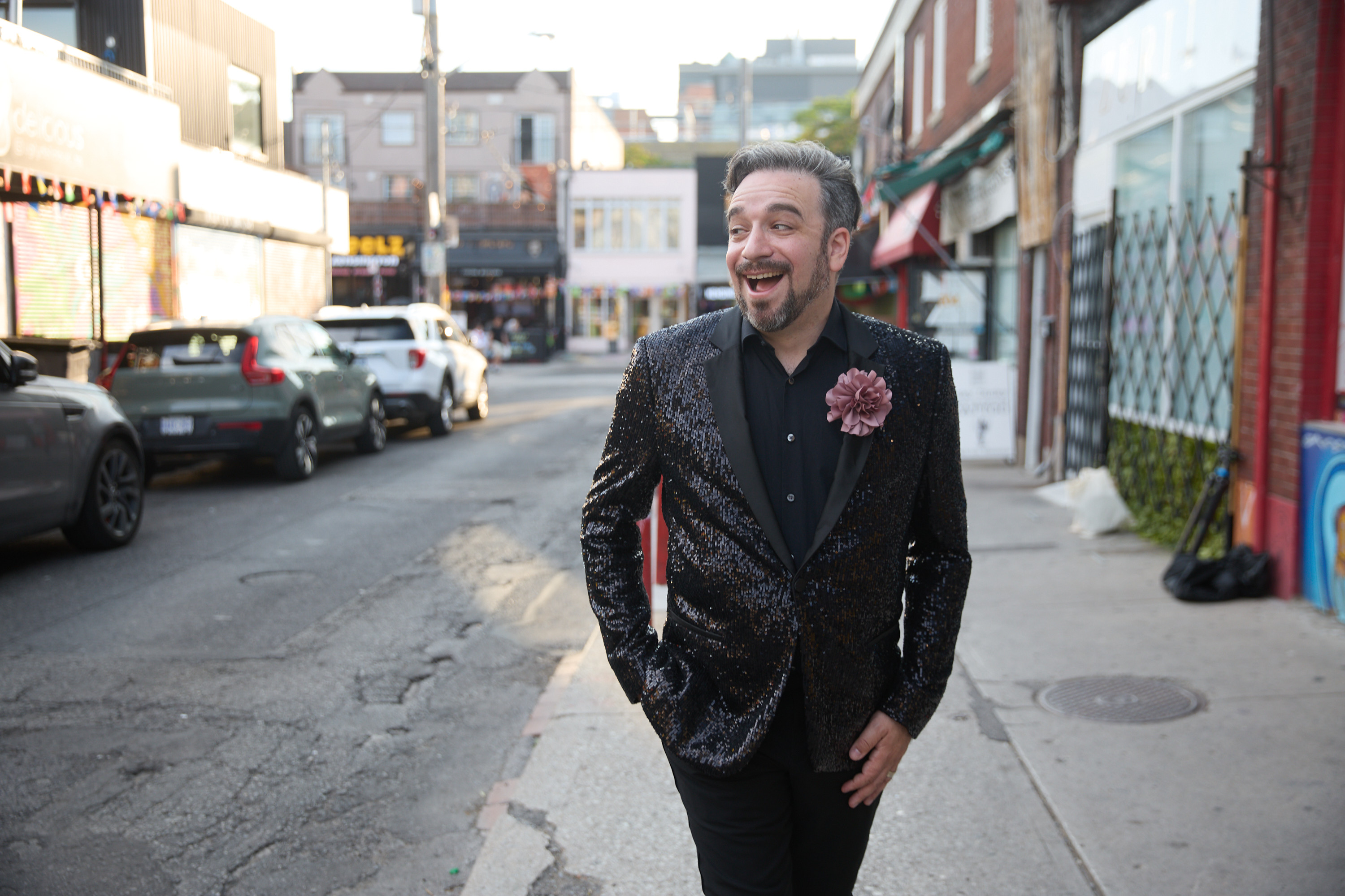 On a daylit city street, a man dressed in a black, sparkly suit and pink carnation struts with a wide smile on his face