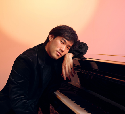 Bruce Liu sits by a piano. Soft pink light flickers over him.
