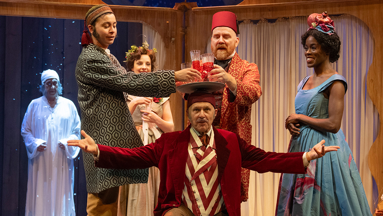 Two actors grab drinks from a platter balanced on a third actor's head as two more look on. Scrooge watches from upstage