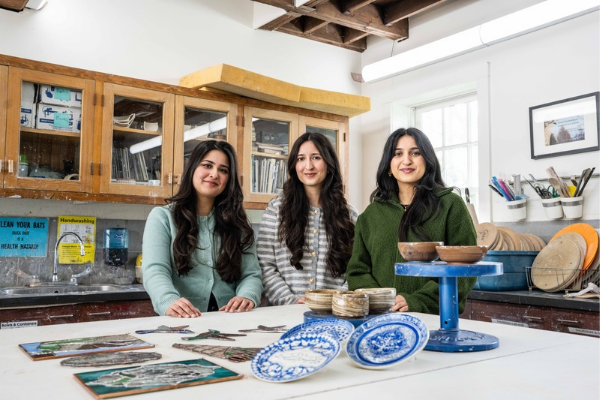 Three figures stand in a ceramic studio with their completed ceramic pieces in front of them on a table top.