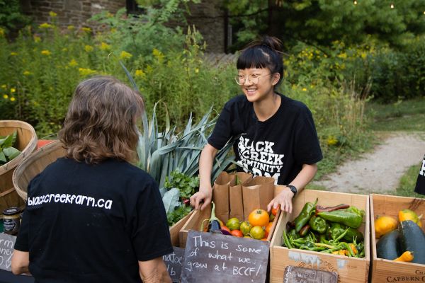 two staff at the booth with fresh seasonal produce in boxes and baskets. Asian woman smiling. Other facing back.