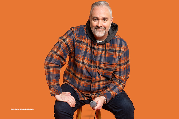 A portrait of a smiling, grey-bearded man in a plaid shirt and hooded jacket holding a microphone against bright orange.