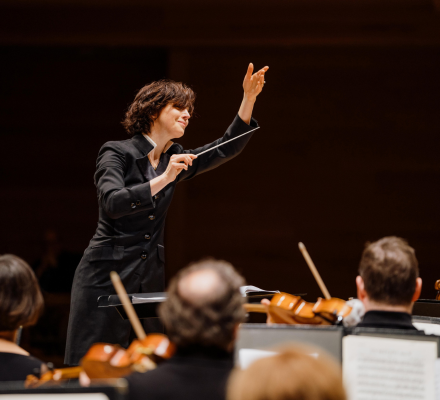 The conductor Tania Miller leads an orchestra with her hand up, holding her baton.