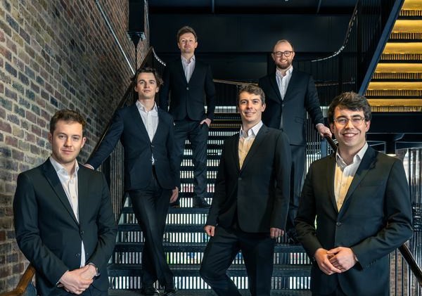A group of six men wearing suits stand together on a staircase.