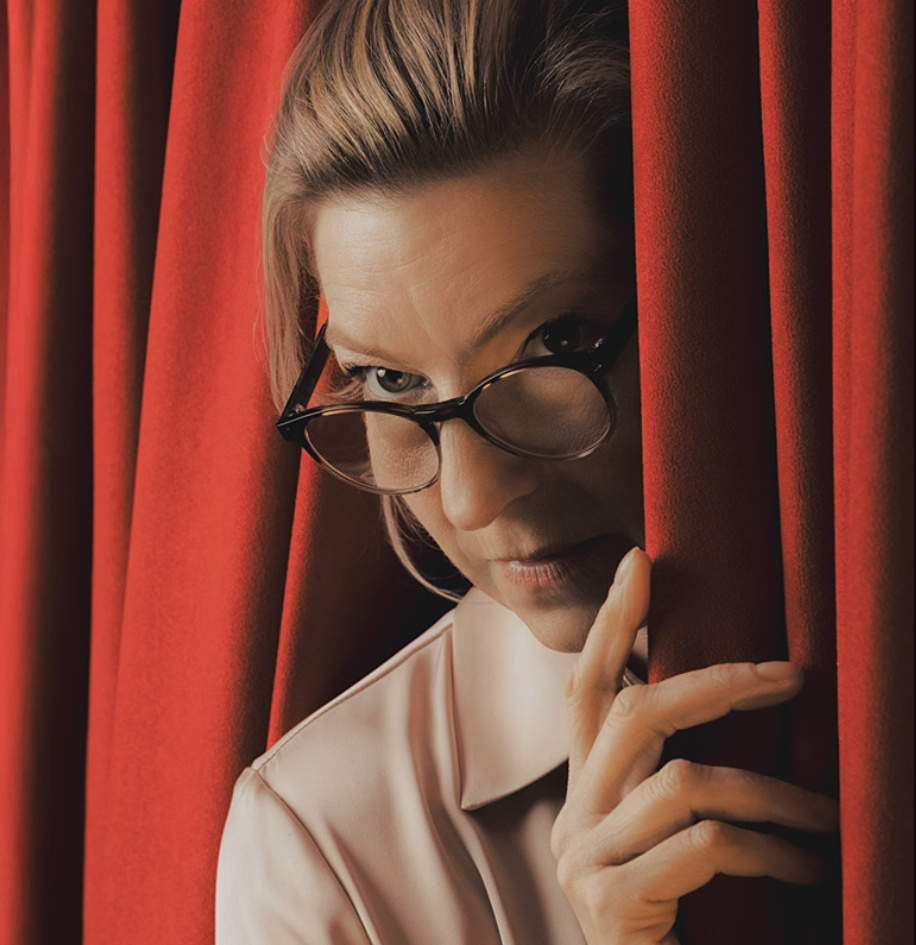 White Woman in glasses peers from behind a dark red stage curtain