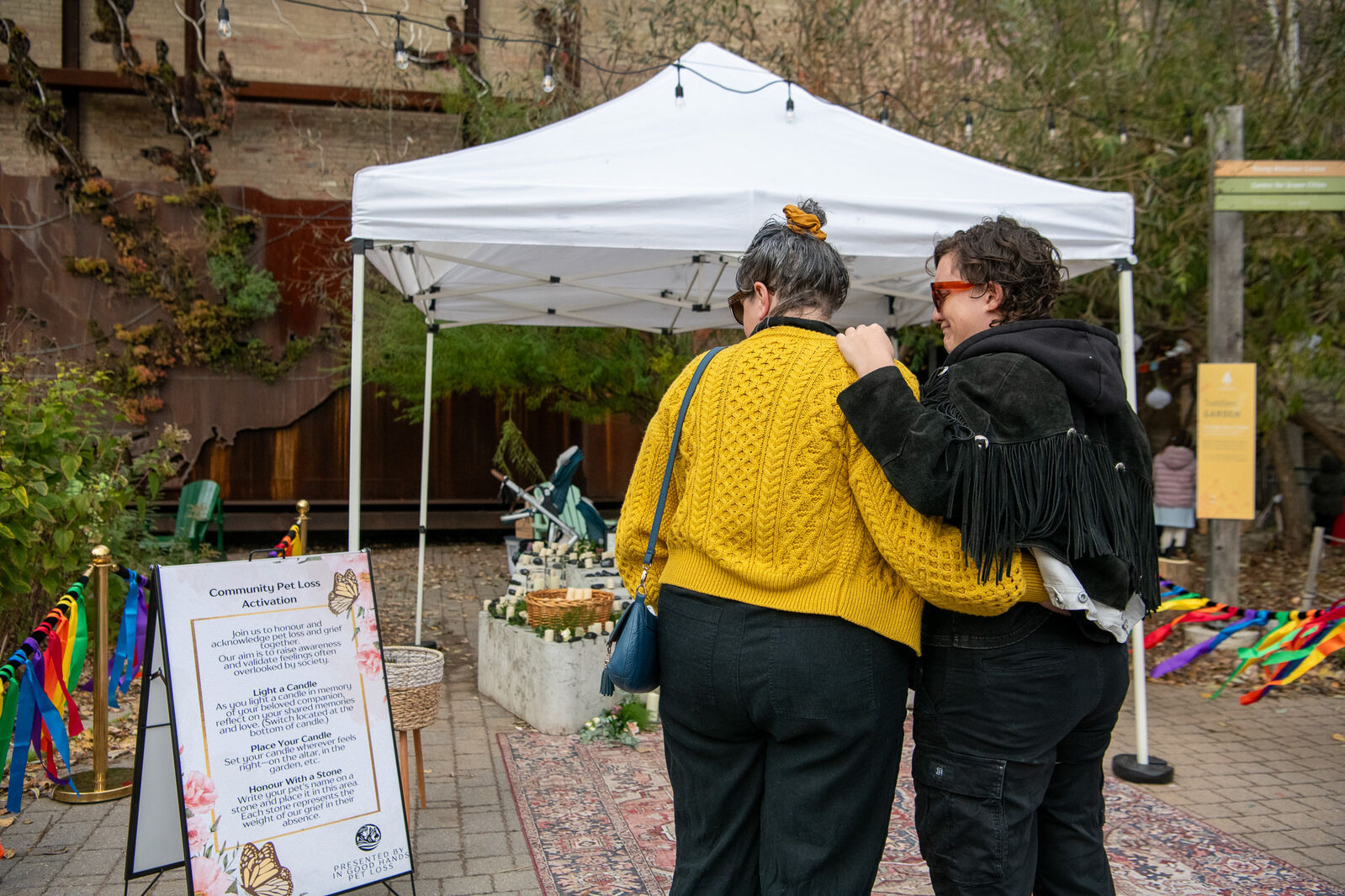 Outdoors, under a white tent, a pet memorial is watched by a woman in a yellow sweater and a woman in black, hugging