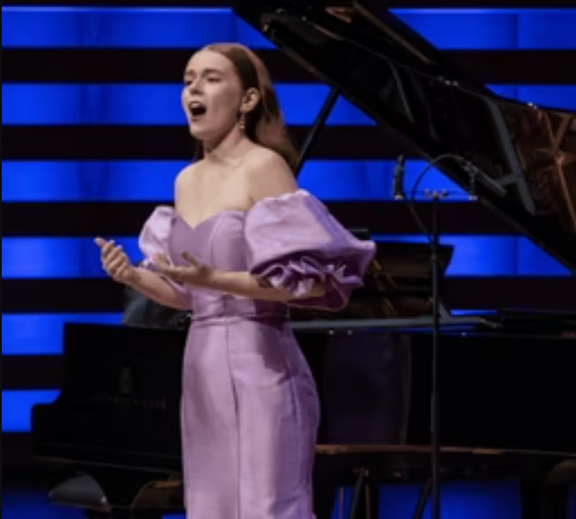 A woman wearing a lilac colour evening dress sings in front of a grand piano.