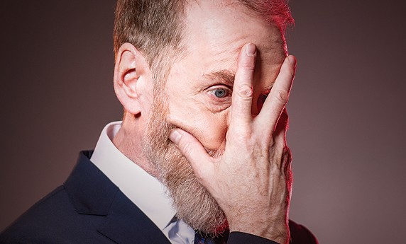 A close-up profile portrait of a bearded man in a suit, covering his face with his hand while peering through his finger