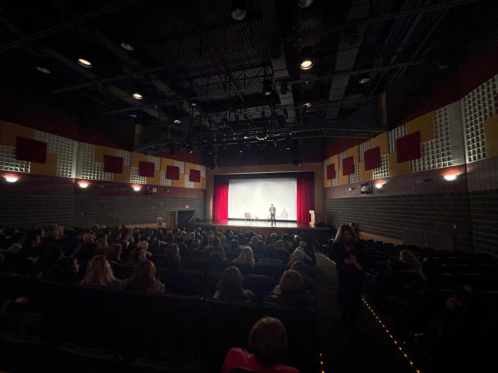 Audience seated in the Hamazkayin Theatre as a speaker addresses the crowd onstage in front of a projection screen.