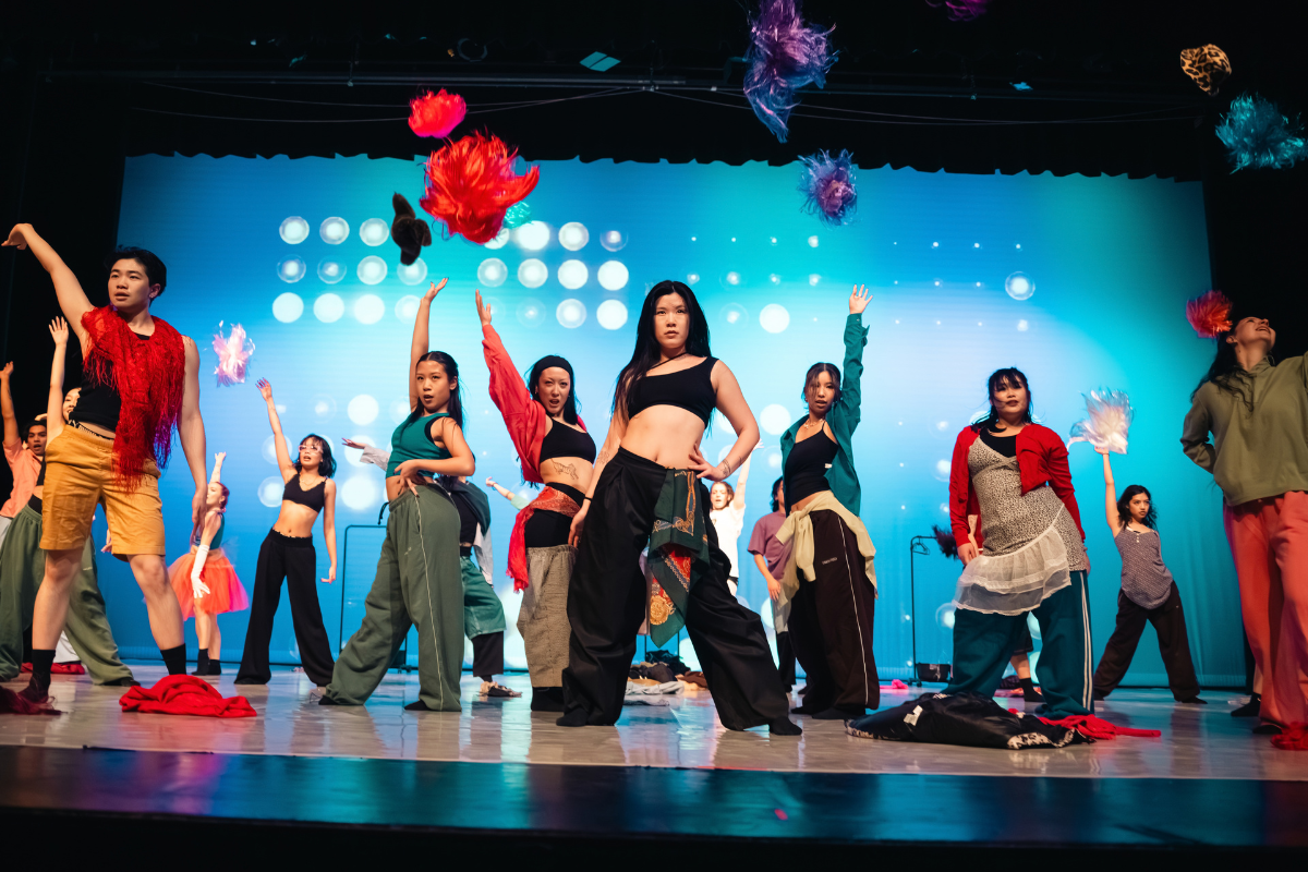 Dancers on stage in bright colourful multi-layers clothes throwing wigs up in the air.