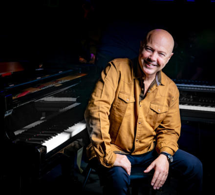 John Beasley wears a yellow sweater and sits at the piano.