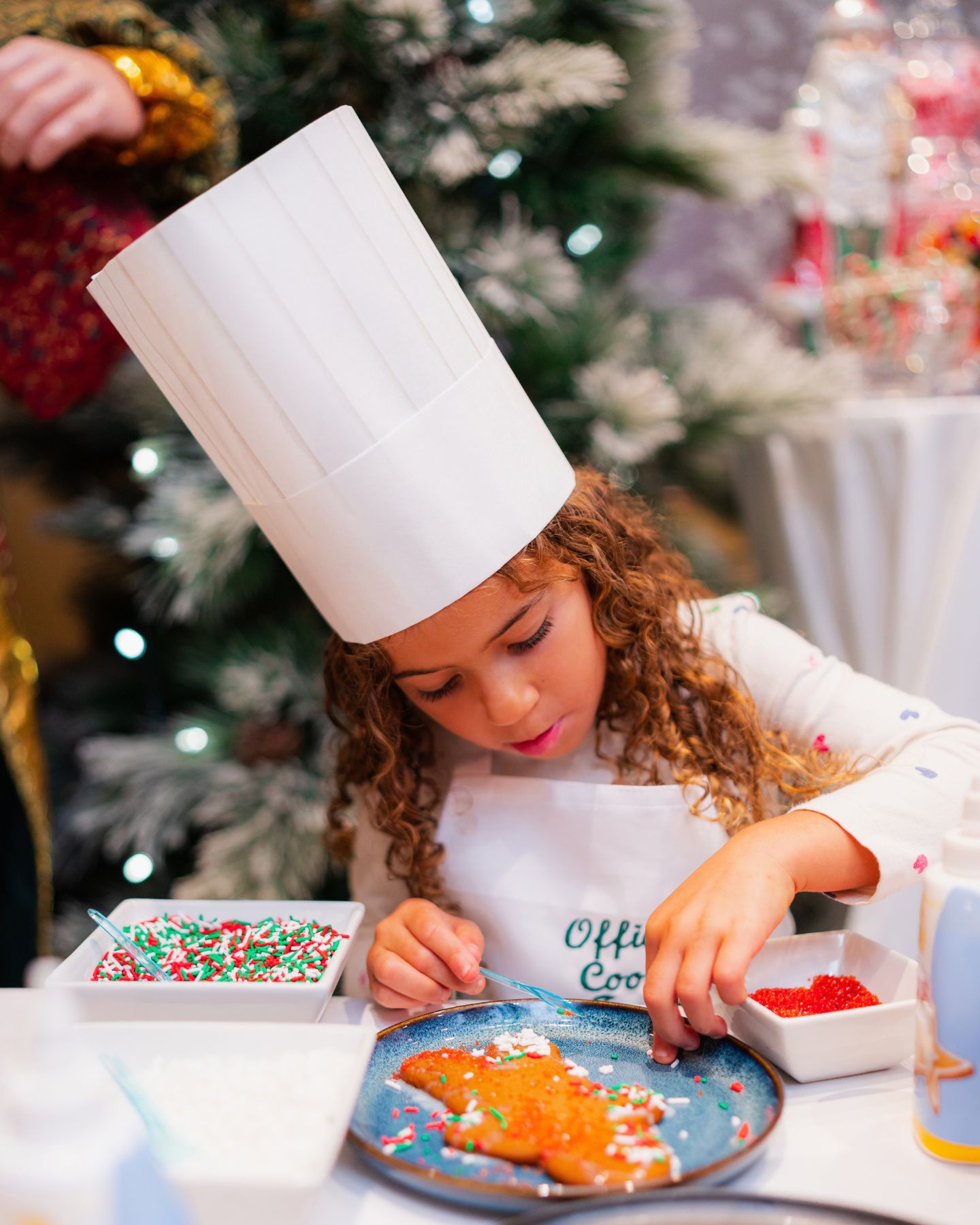 With a backdrop of a Christmas tree and decorations, a kid wearing a chef's hat and apron, decorates a festive cookie
