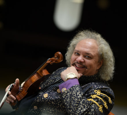 Roby Lakatos, smiles. He has curly grey hair and wears a sparkly black suit with purple details. He holds his violin.