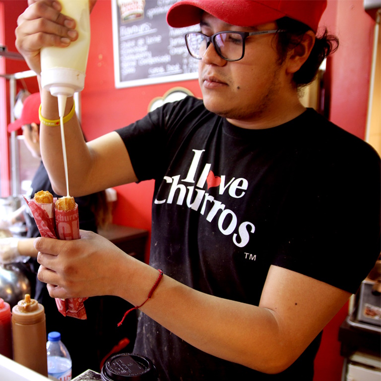 Inside a restaurant, an employee wearing a black shirt that says I Love Churros drizzles sauce over two churros.