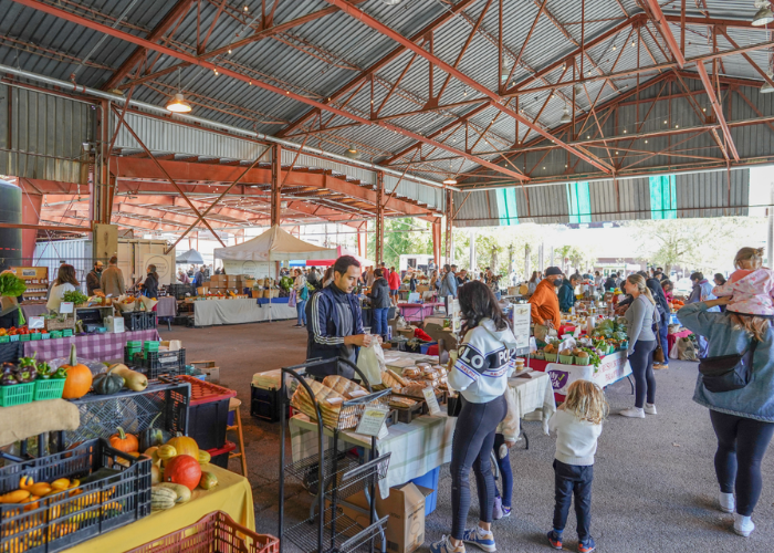 Shoppers browsing market stalls under a pavilion roof.