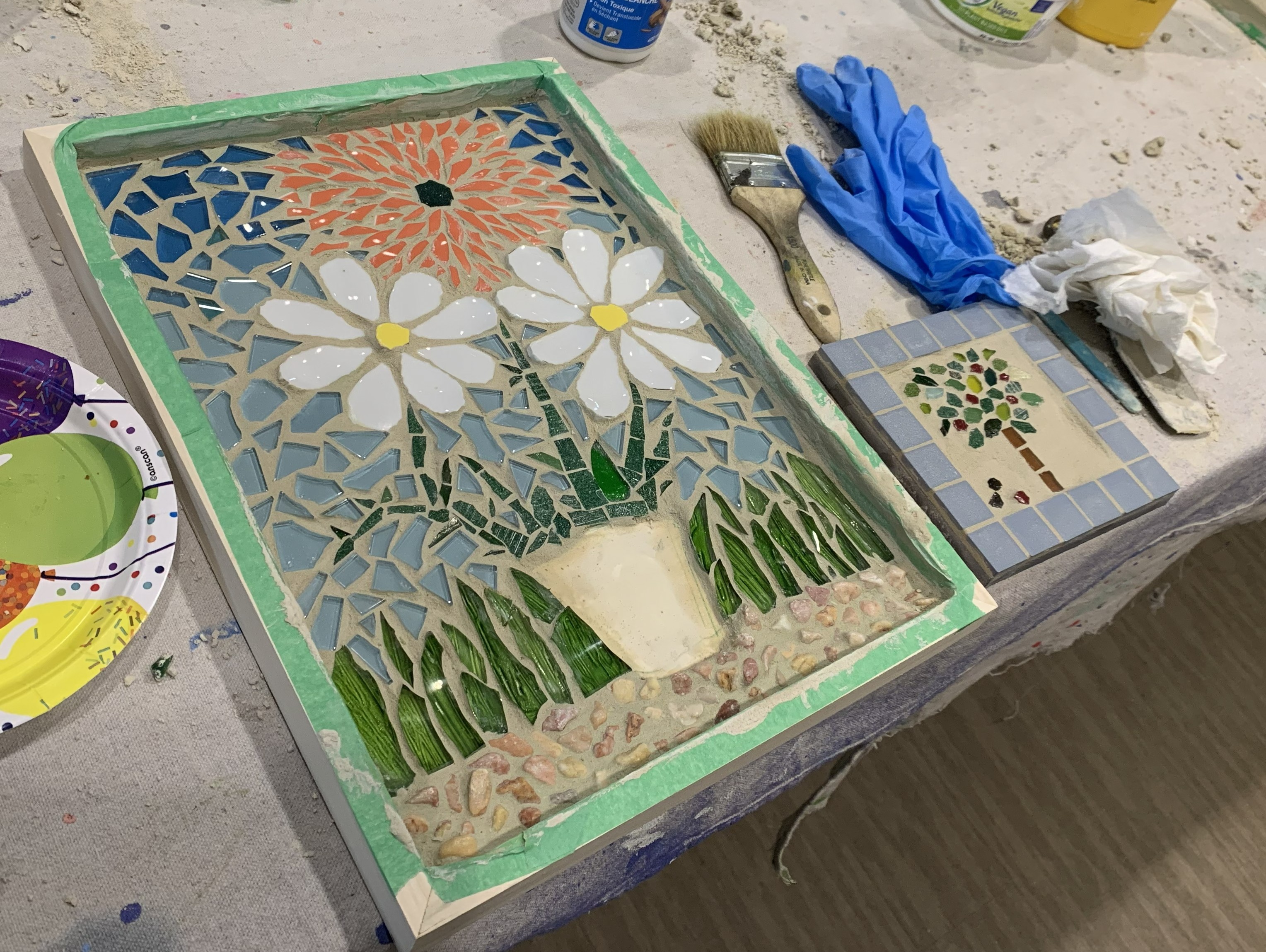 An unfinished mosaic piece of flowers in a flower pot on a table with gloves, paints and glue.