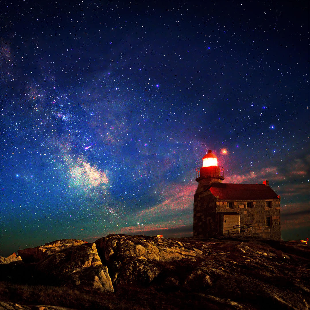 Stone lighthouse sat upon rocks with northern lights across the night sky