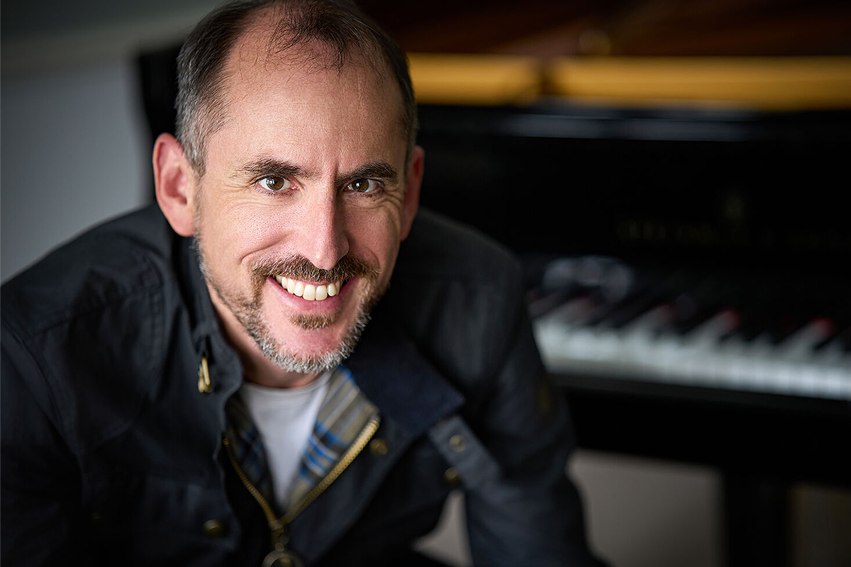 Smiling man in a black shirt, with a piano in the background