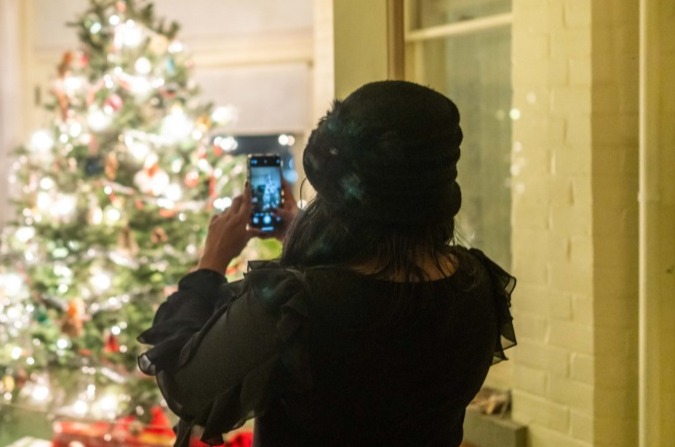 A woman takes a cell phone photo of an illuminated Christmas tree.