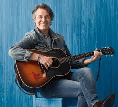 Jim Cuddy holds a guitar in his lap. He wears a jean jacket and jeans and sits in front of a blue background.