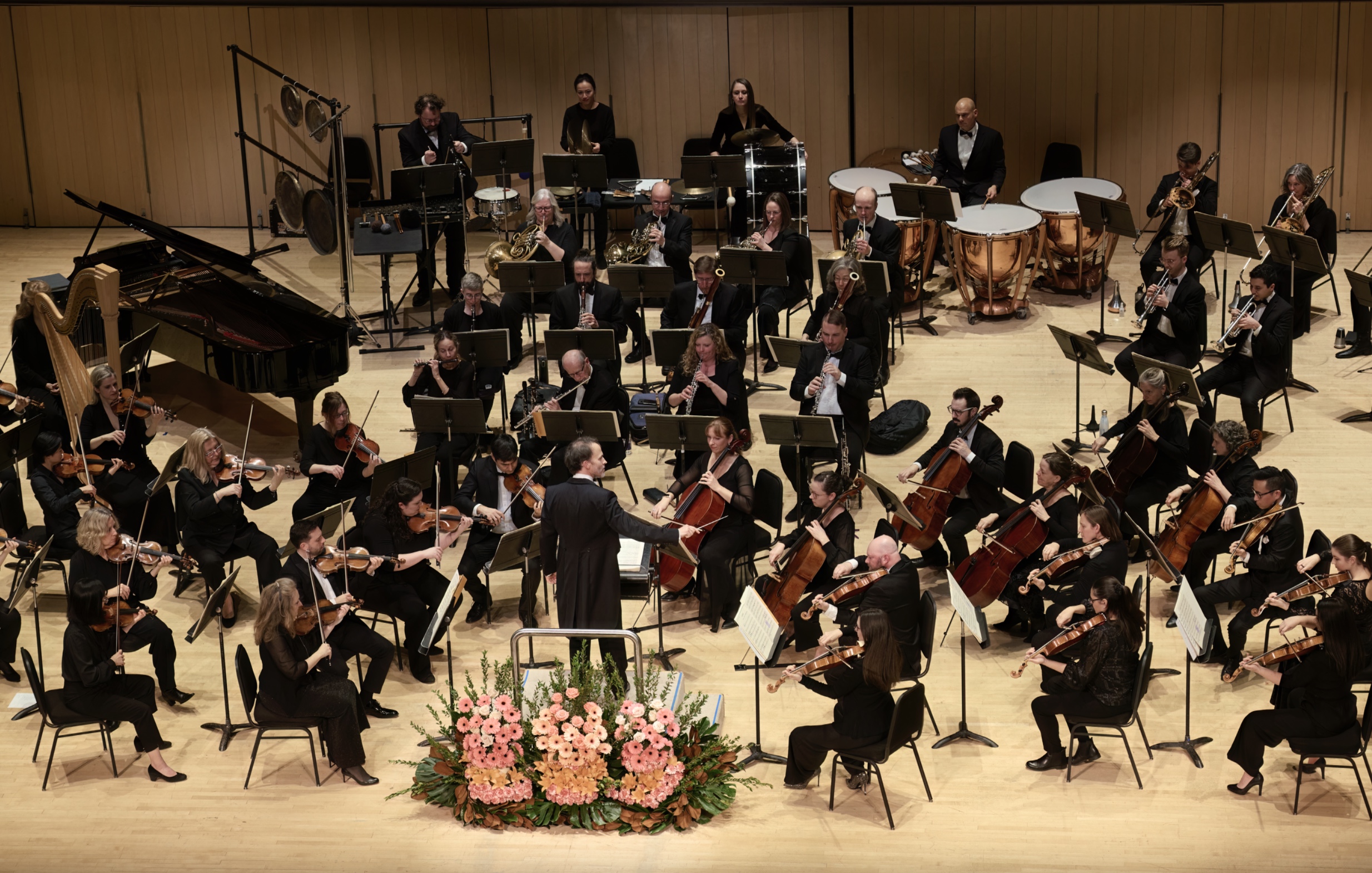 A full orchestra performs onstage under the direction of a conductor, surrounded by elegant floral arrangements.
