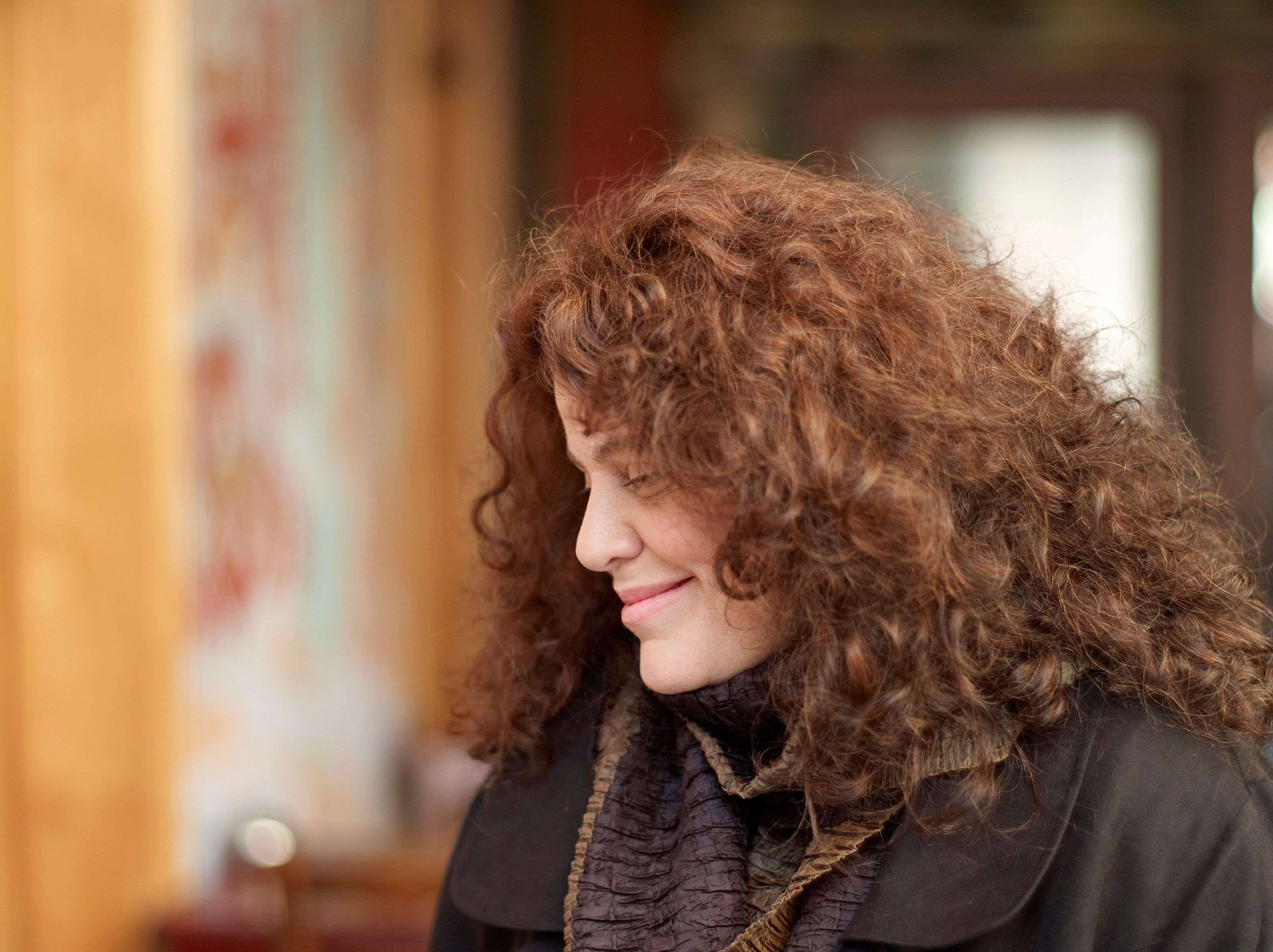 Person with curly hair wearing layered dark clothing, standing indoors with wooden furniture and soft lighting.