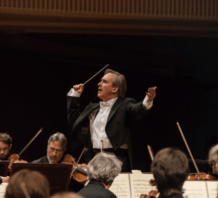 James Conlon performs on stage. He wears a full suit, with his hand raised with a baton.