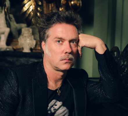 Rufus Wainwright props his head up with his arm. He wears a black suit jacket.